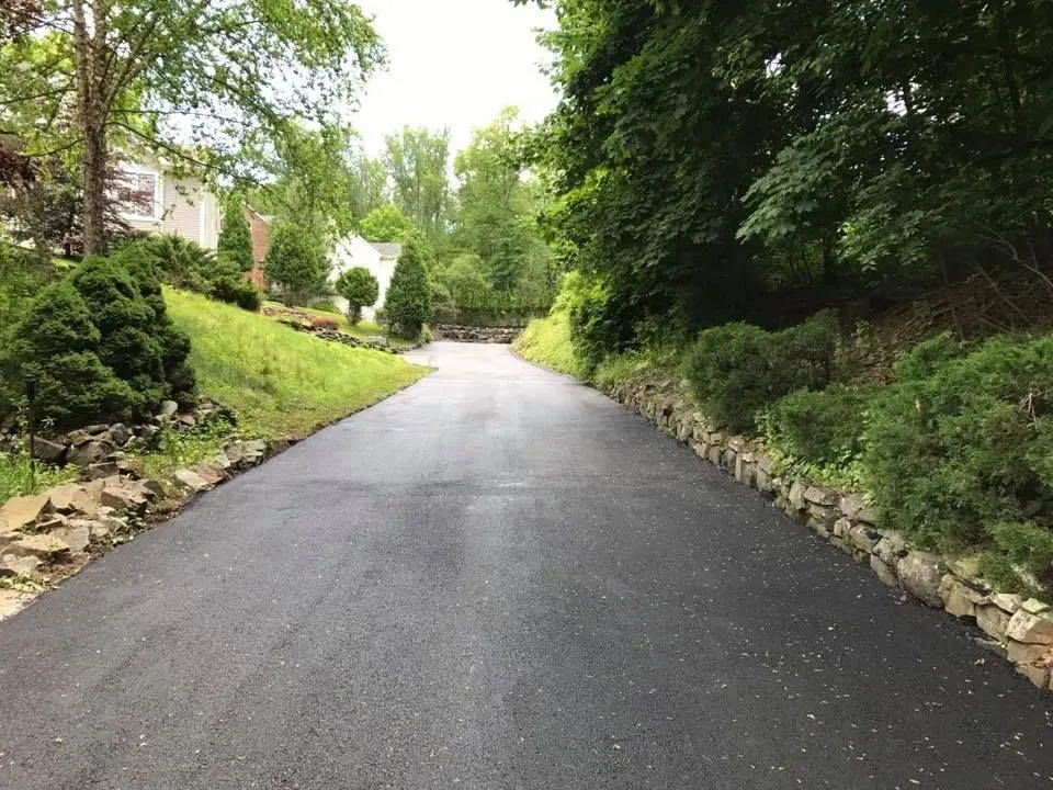 Paved driveway slopes uphill, bordered by greenery and stone retaining walls.