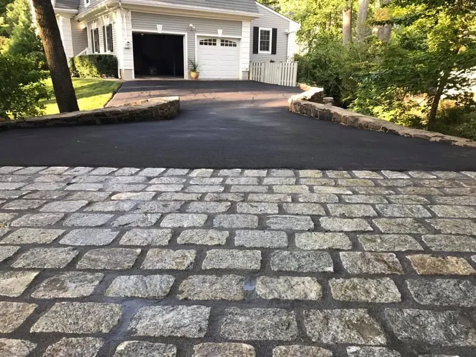 Cobblestone driveway leading to a black asphalt driveway with a house and garage in the background.
