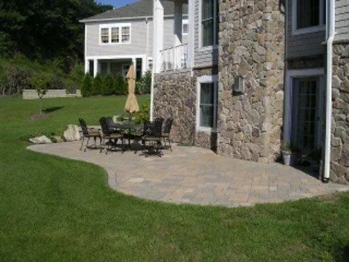 Stone patio with table and chairs next to house with stone and siding facade, green lawn.