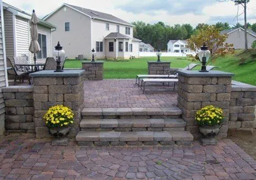 Stone patio with steps, pillars, and a brick pattern, with flowerpots and a house in the background.