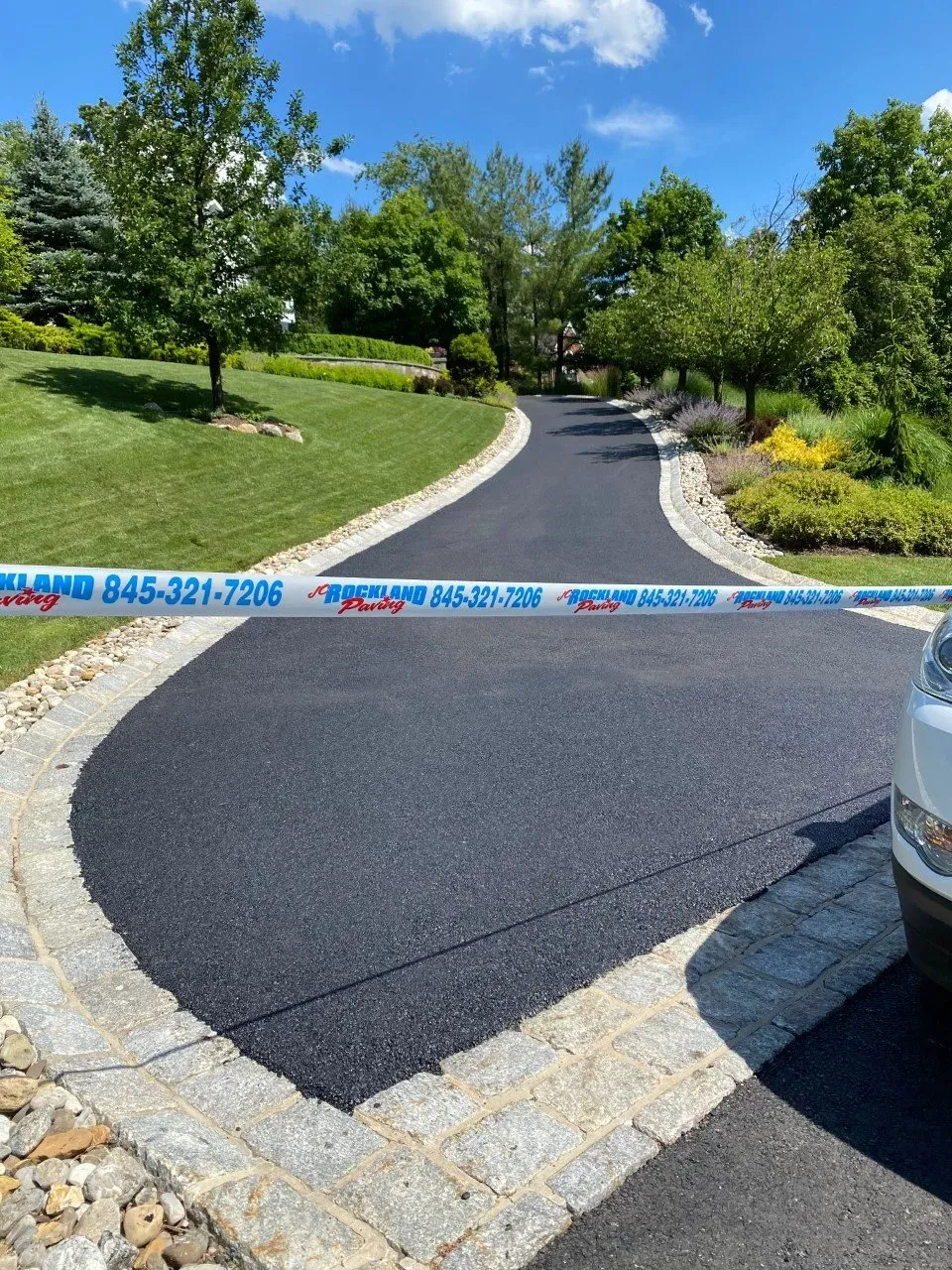 Freshly paved asphalt driveway with cobblestone edging, blocked by blue caution tape under a sunny sky.