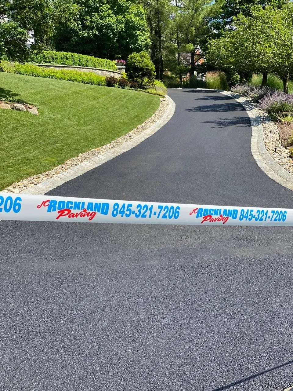Freshly paved black driveway with caution tape in front. Green lawn on left, garden on the right.