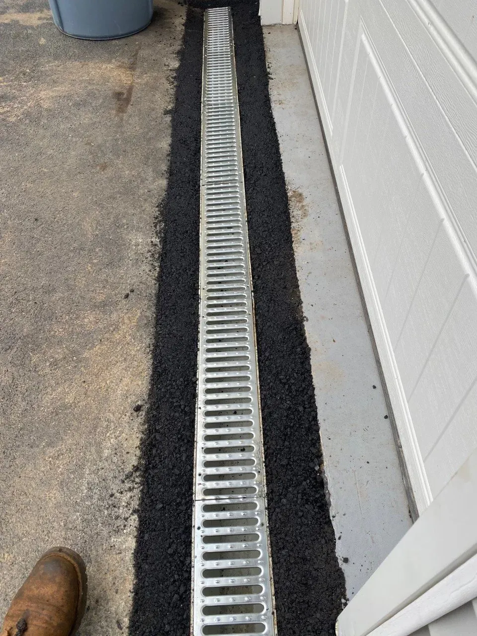 A linear trench drain with a metal grate installed in asphalt beside a white garage door.