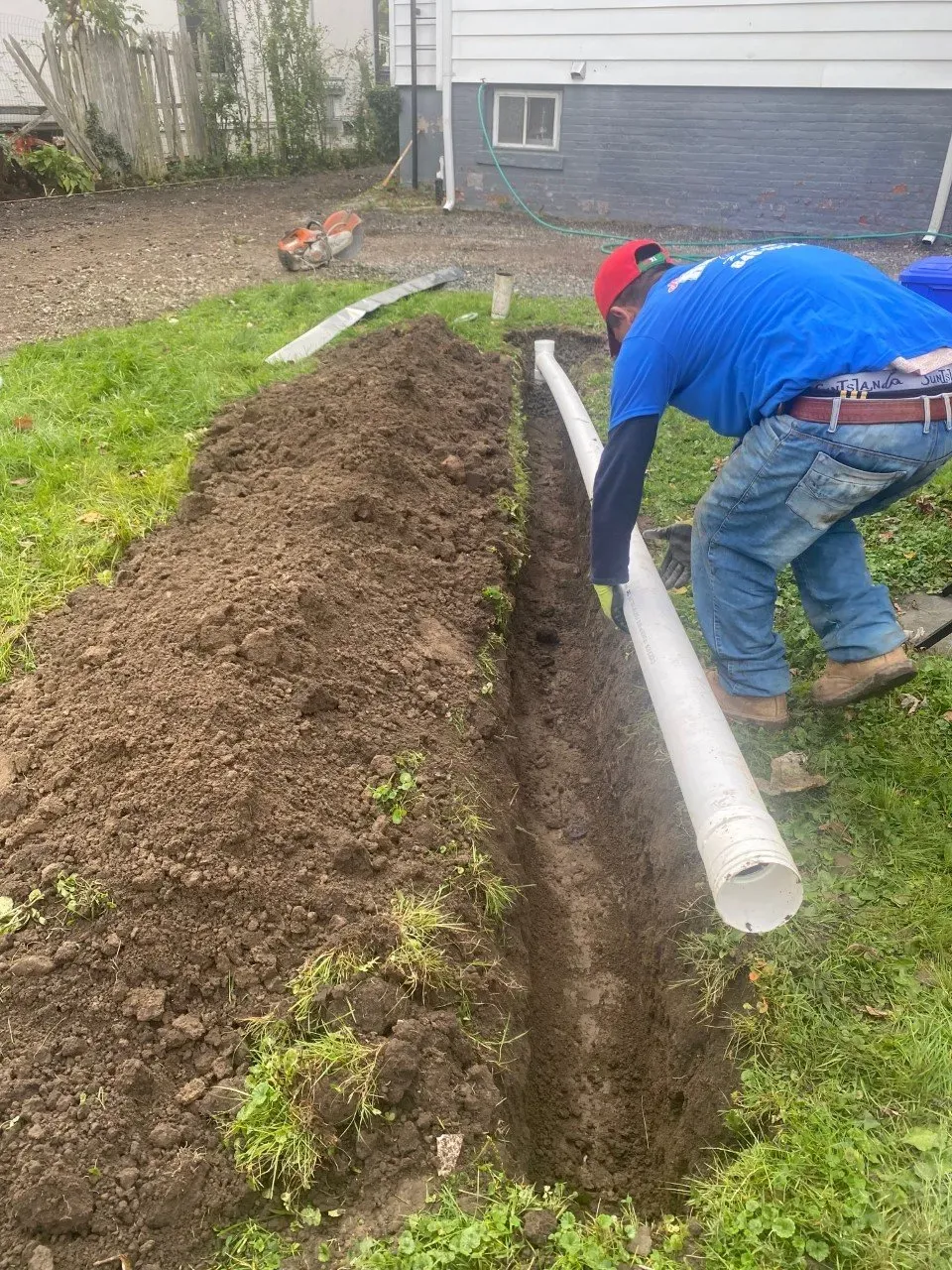 Man installing white drainage pipes in a trench in a grassy backyard, near a building.