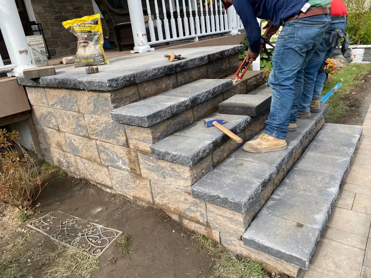 Workers building stone steps on a porch, using tools. The steps are gray and the wall is brown.