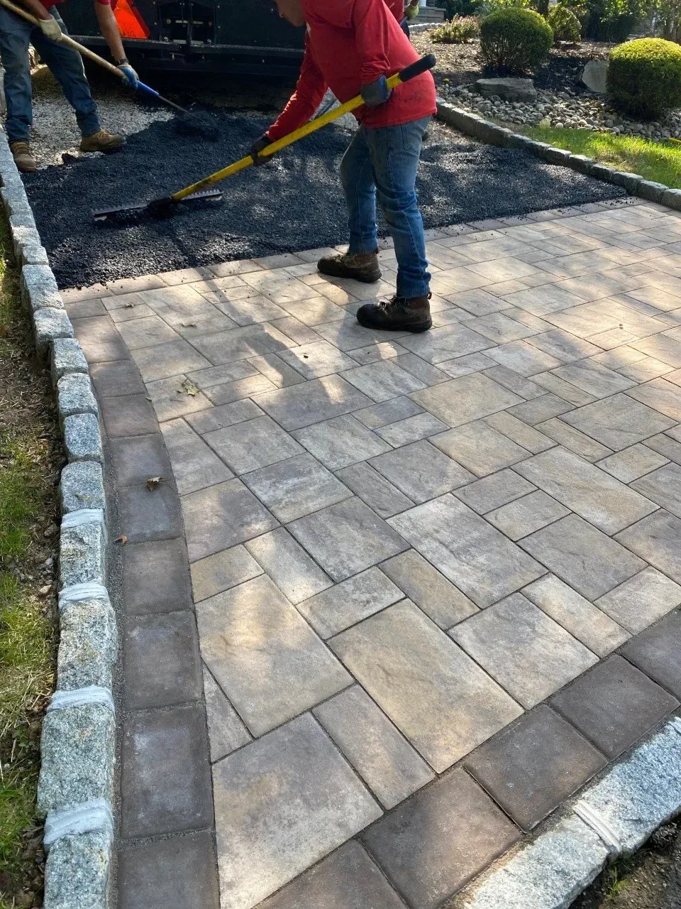 Workers paving a walkway: asphalt being laid next to patterned pavers, bordered with stone.
