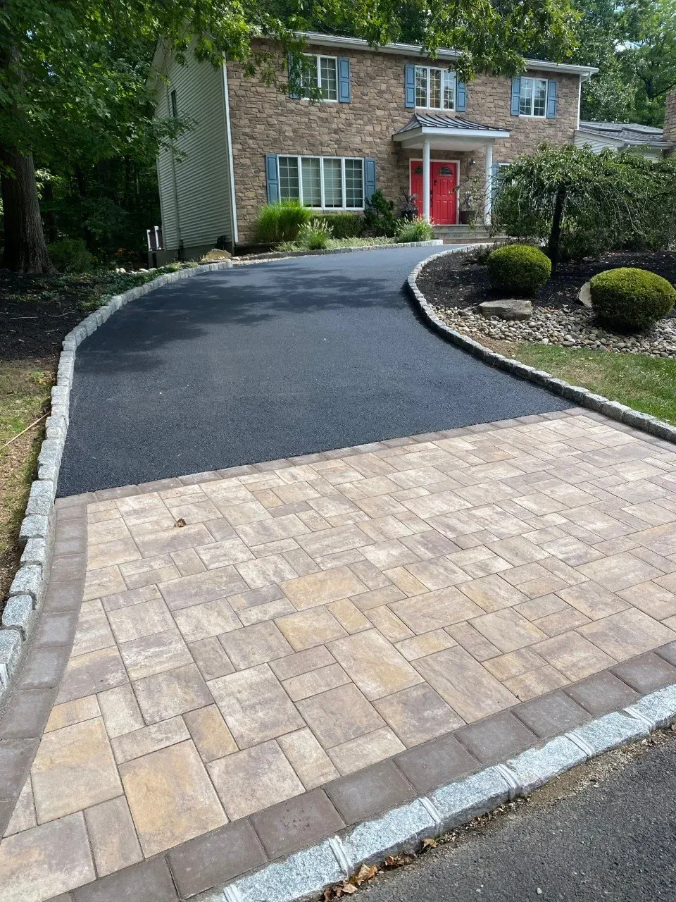 Driveway with brick pavers and asphalt leading to a two-story brick house with a red door.