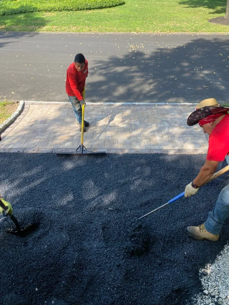 Two workers laying asphalt on a driveway, one using a rake, the other shoveling.
