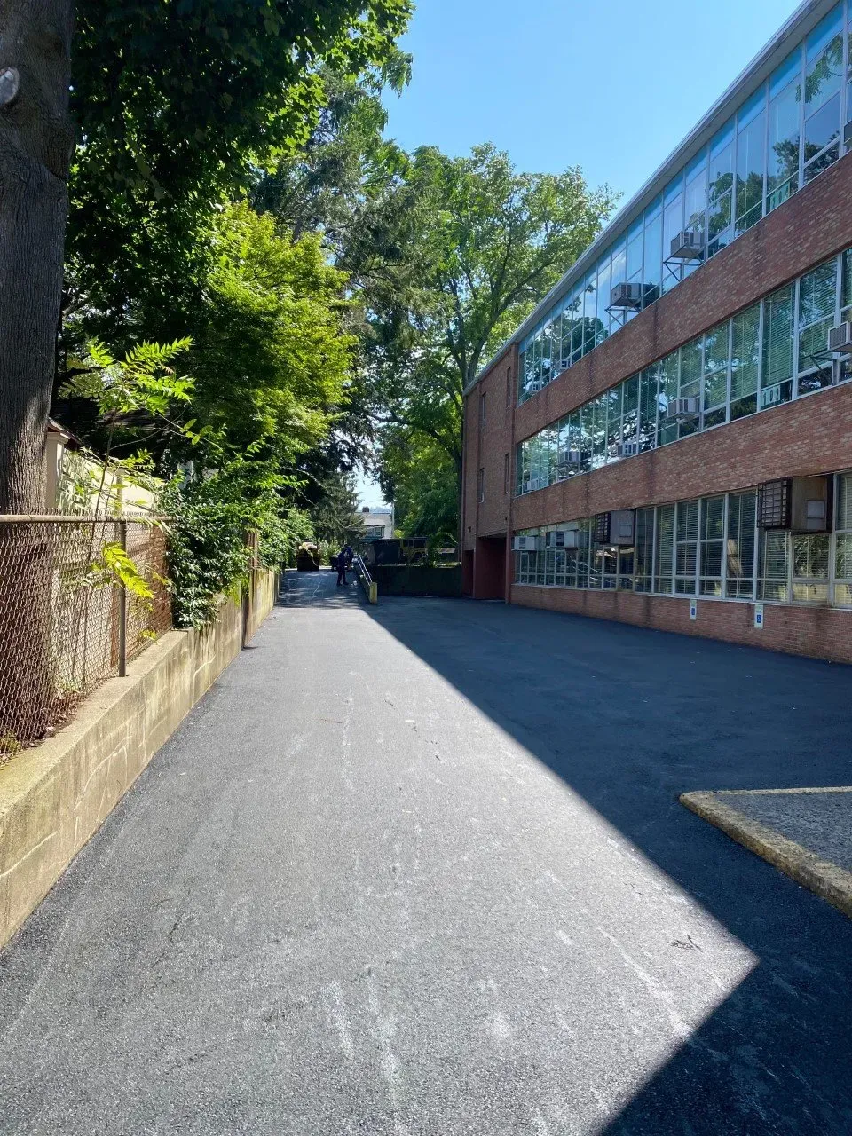 Asphalt walkway alongside a brick building with large windows and a chain-link fence, under a sunny sky.