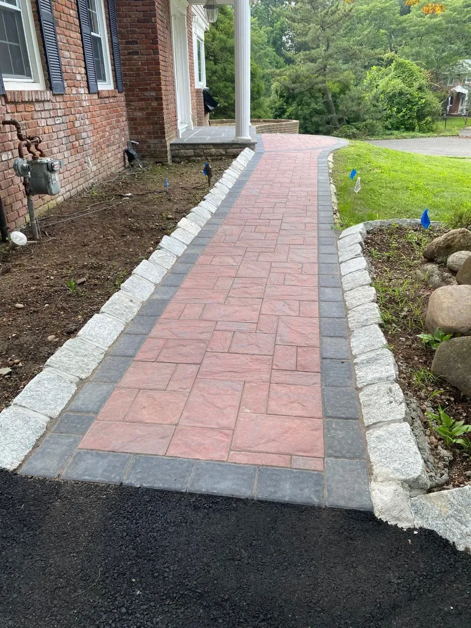 Brick pathway with gray border, leading to a home's porch, bordered by landscaping and fresh asphalt.