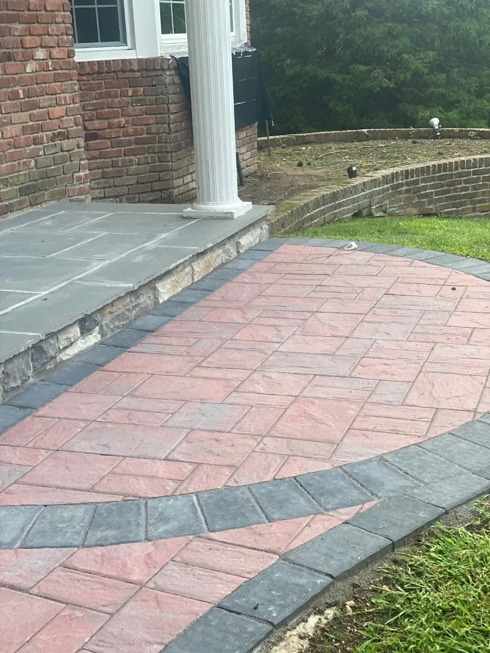 Brick and stone patio with a curved border, next to a brick wall and a white column.