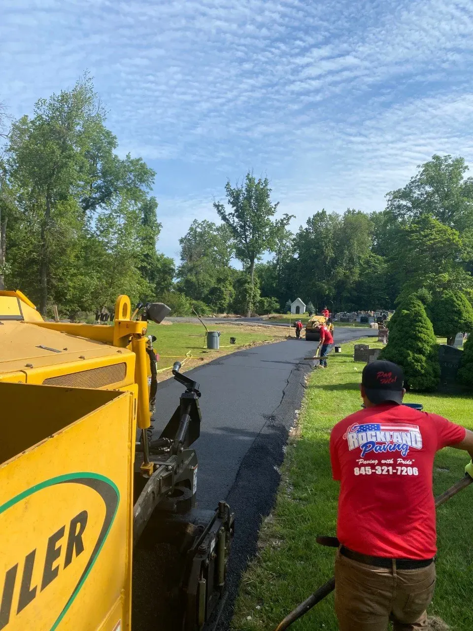 Asphalt paving in a cemetery on a sunny day. A worker in a red shirt guides paving.