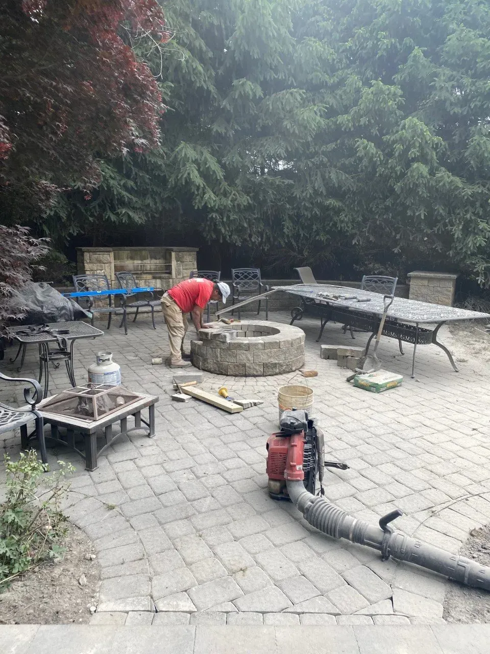 Person working on a stone fire pit in a brick patio, surrounded by outdoor furniture.