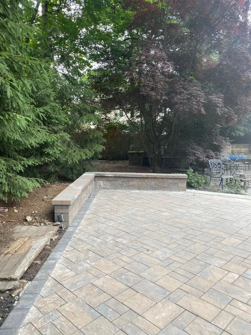Patio with brick pavers and low retaining wall, adjacent to a wooded area with trees.