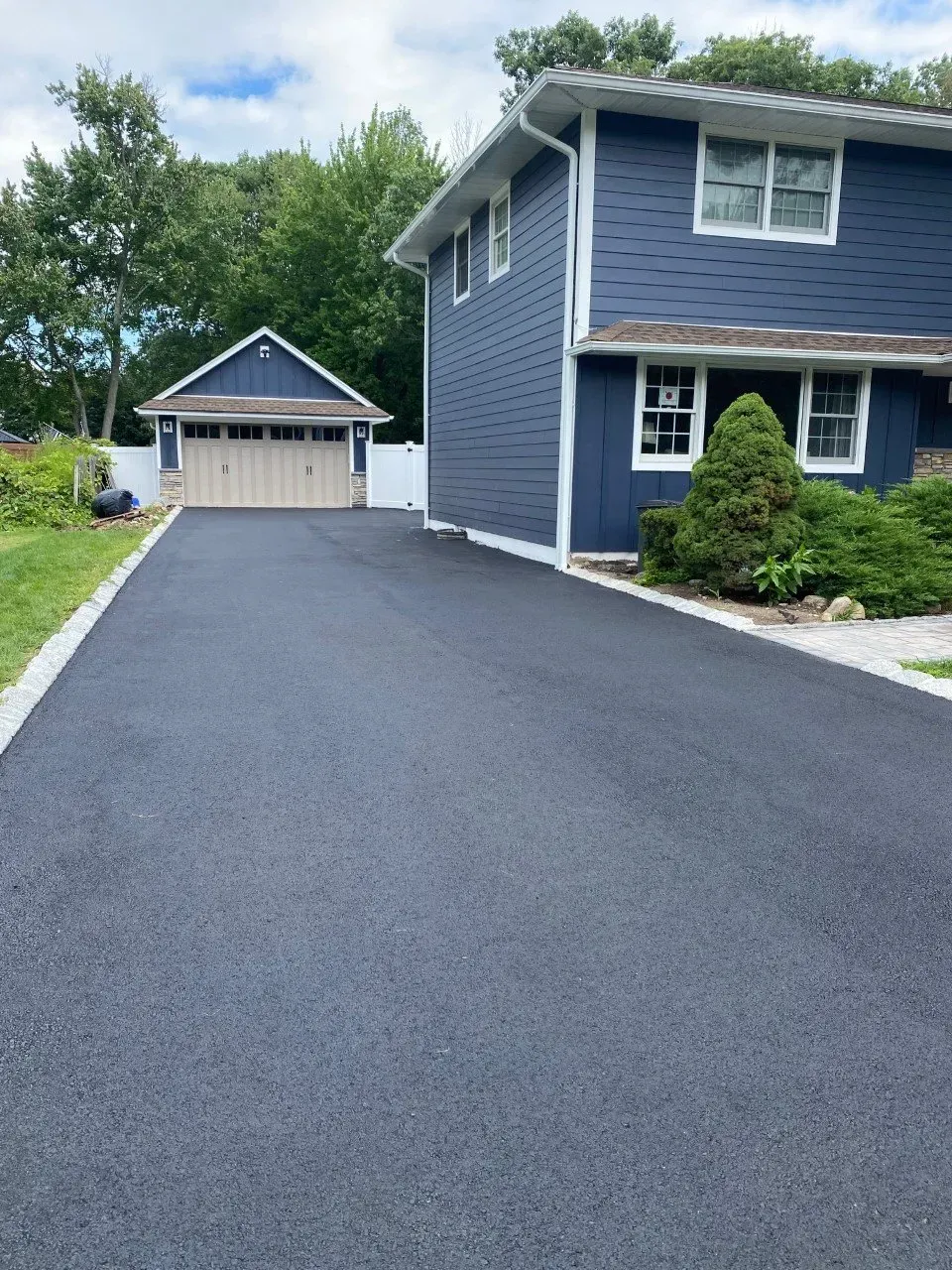 Asphalt driveway leading to a blue two-story house and detached garage with tan doors.