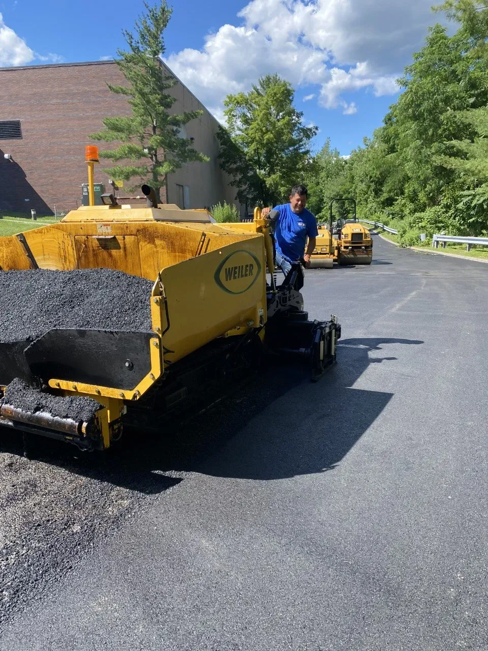 Asphalt paving on a road with a paving machine and worker. Yellow machine, blue shirt. Sunny day.