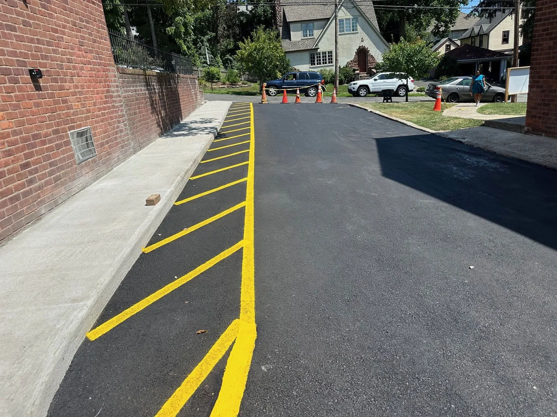 Newly paved asphalt area with yellow diagonal lines, next to a brick wall and a concrete sidewalk.