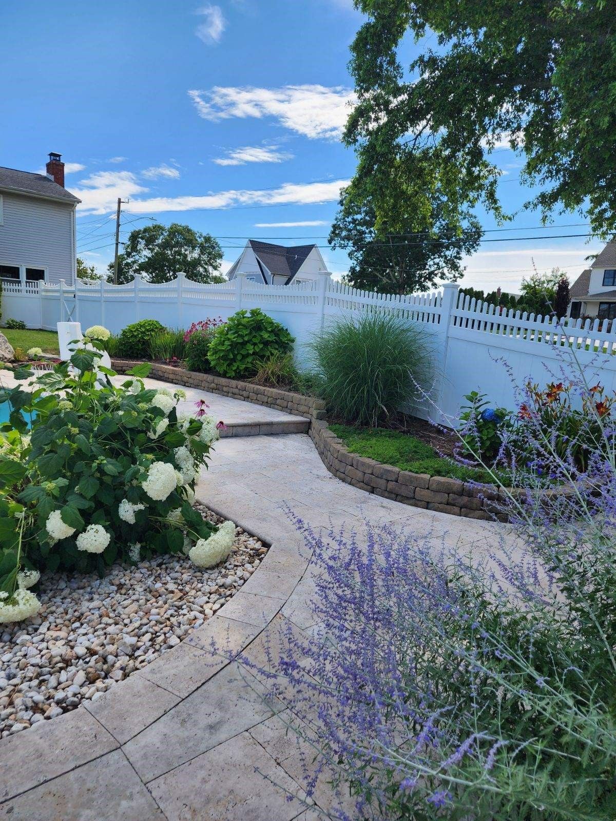 A white fence surrounds a garden with flowers and bushes.