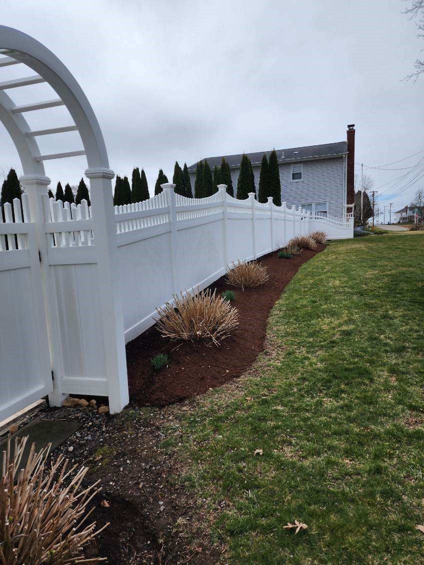 A white fence surrounds a lush green yard with a house in the background.