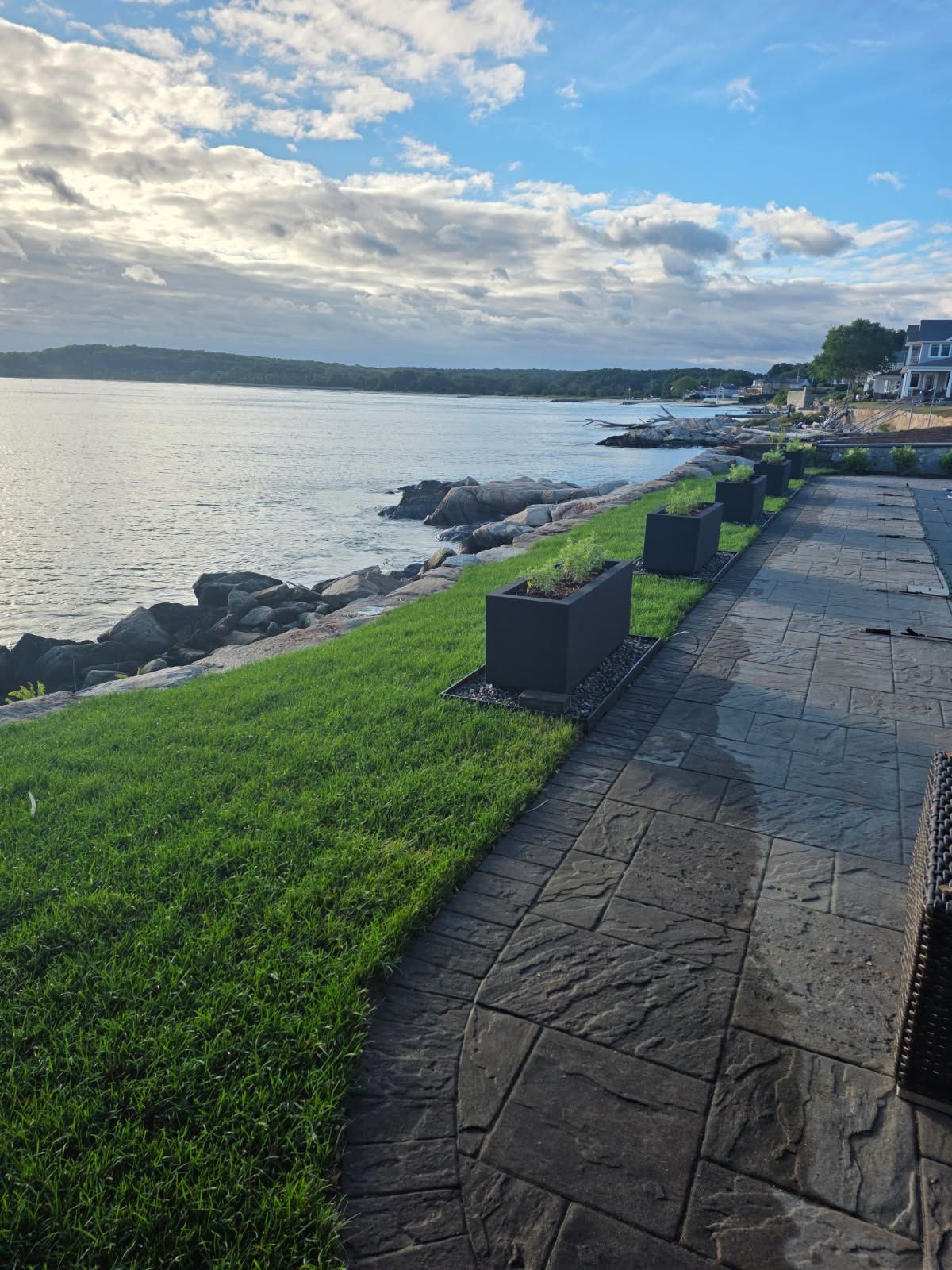 Waterfront scene with green grass, a stone walkway, and calm water under a cloudy sky.