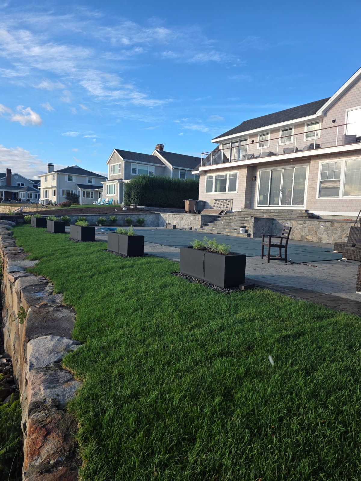 Lush green lawn with dark planters in front of multi-story homes under a blue sky.