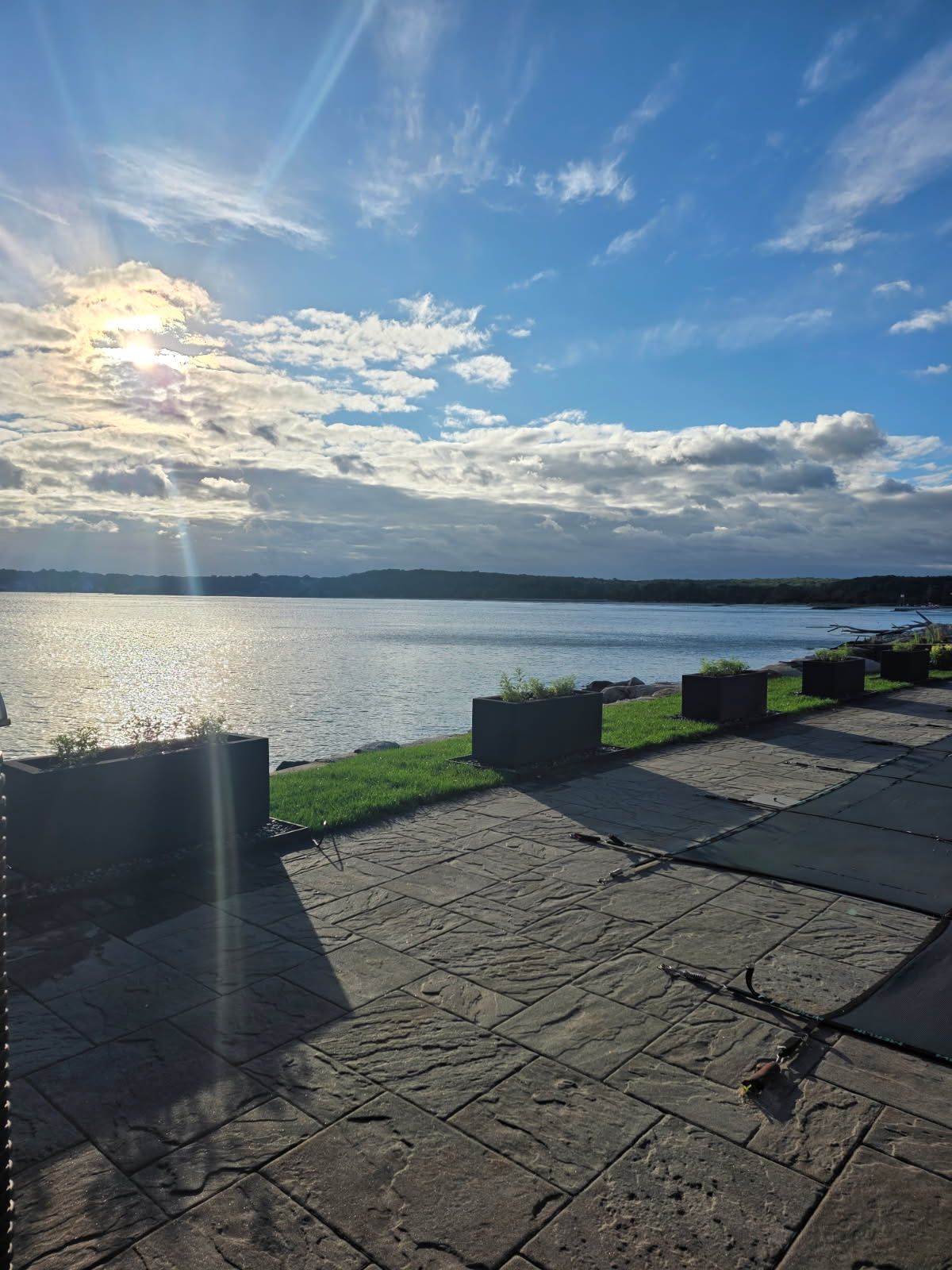 Bright sunlight over water, stone patio in foreground, grassy area with planters. Blue sky with clouds.