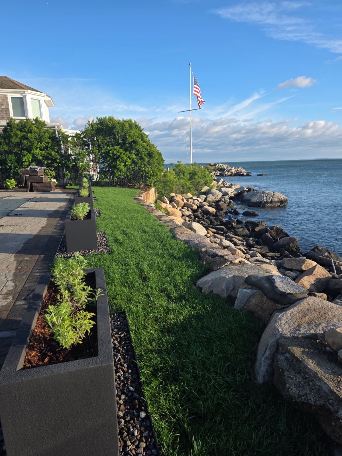 Coastal scene: Lawn, rocks, water, American flag on a pole, dark planters with plants, blue sky.