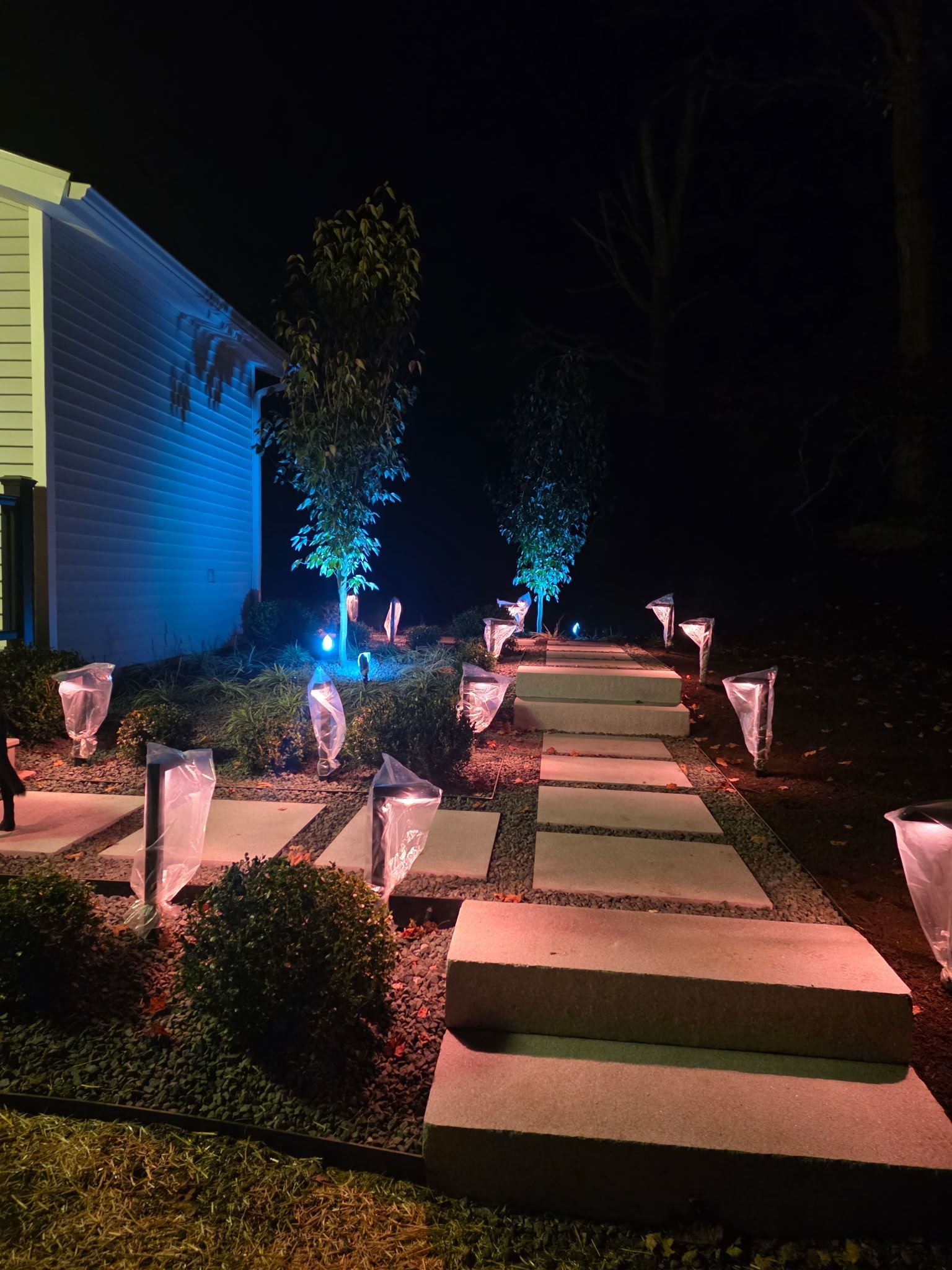A lit pathway with square stepping stones leads to a house at night, lit with colorful garden lights.