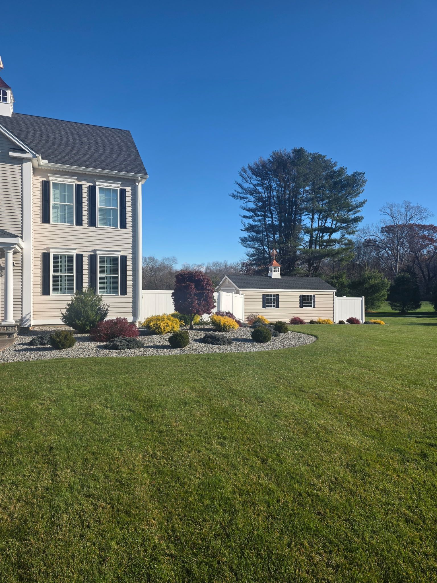 Two-story beige house with black shutters, lush green lawn, small shed, and clear blue sky.