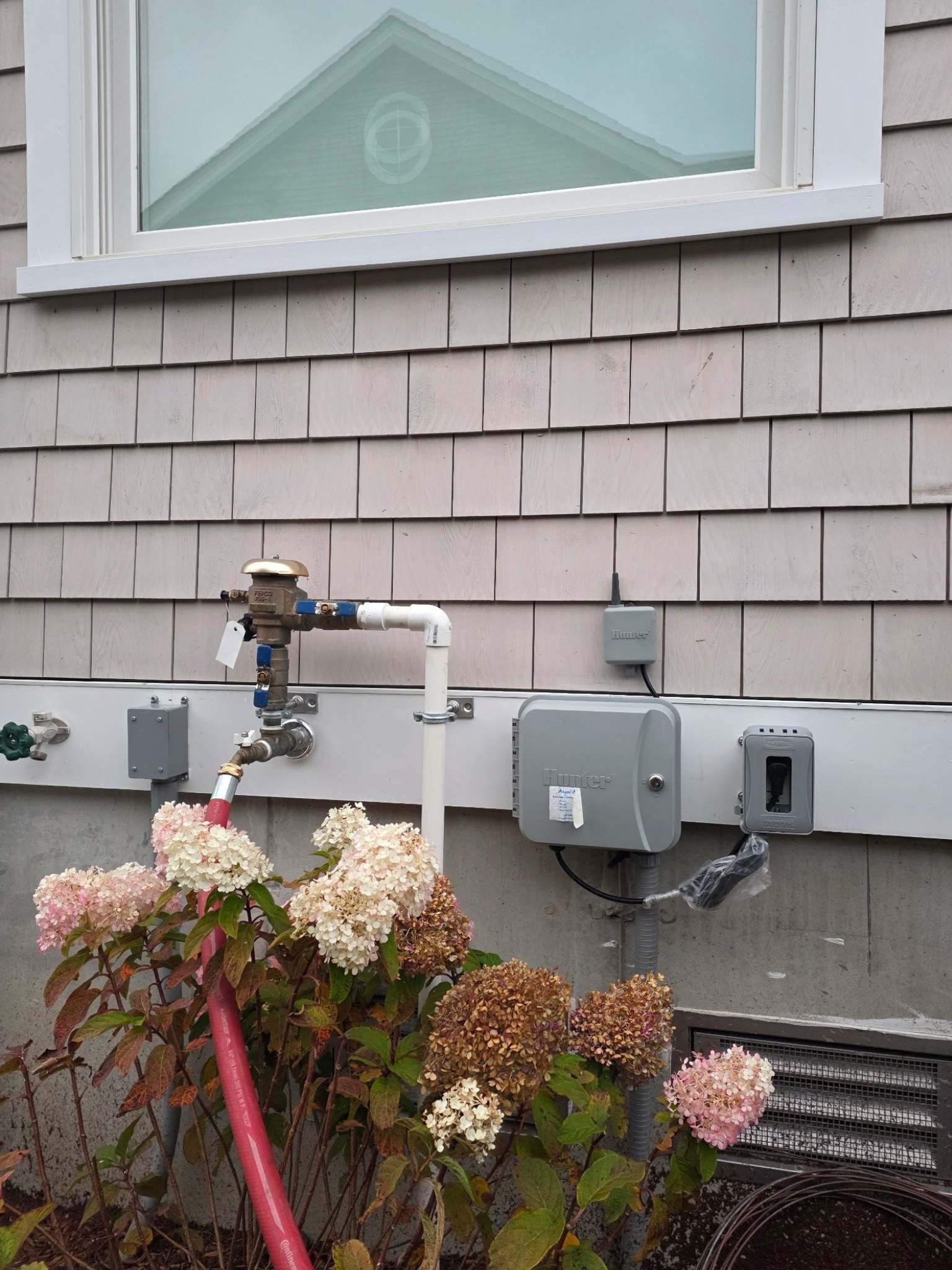 Exterior view of a house with light gray shingles, window, water meter, irrigation system, and pink hydrangea flowers.