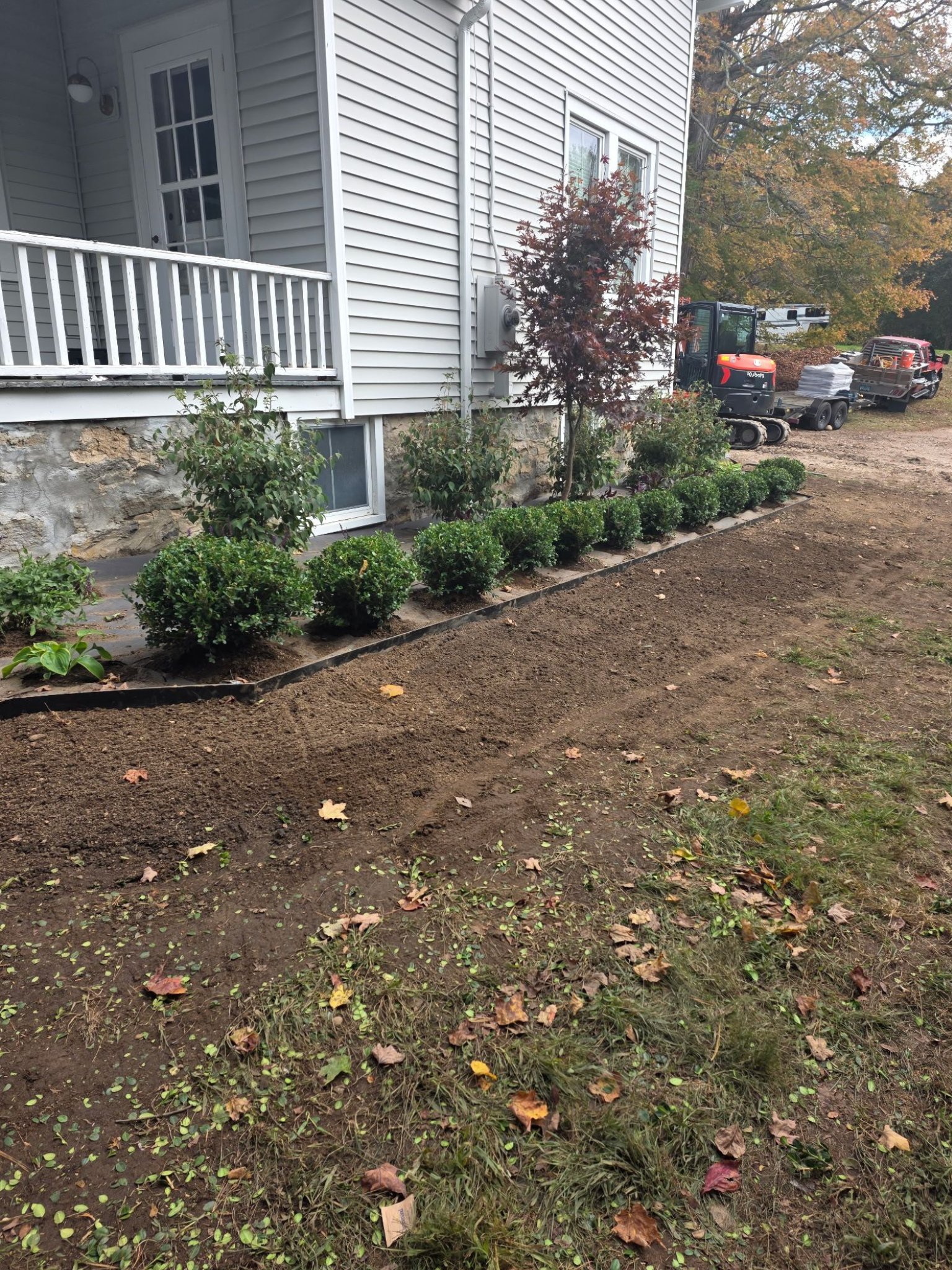 Newly planted shrubs line a flower bed beside a white house with a porch; fall foliage is visible.