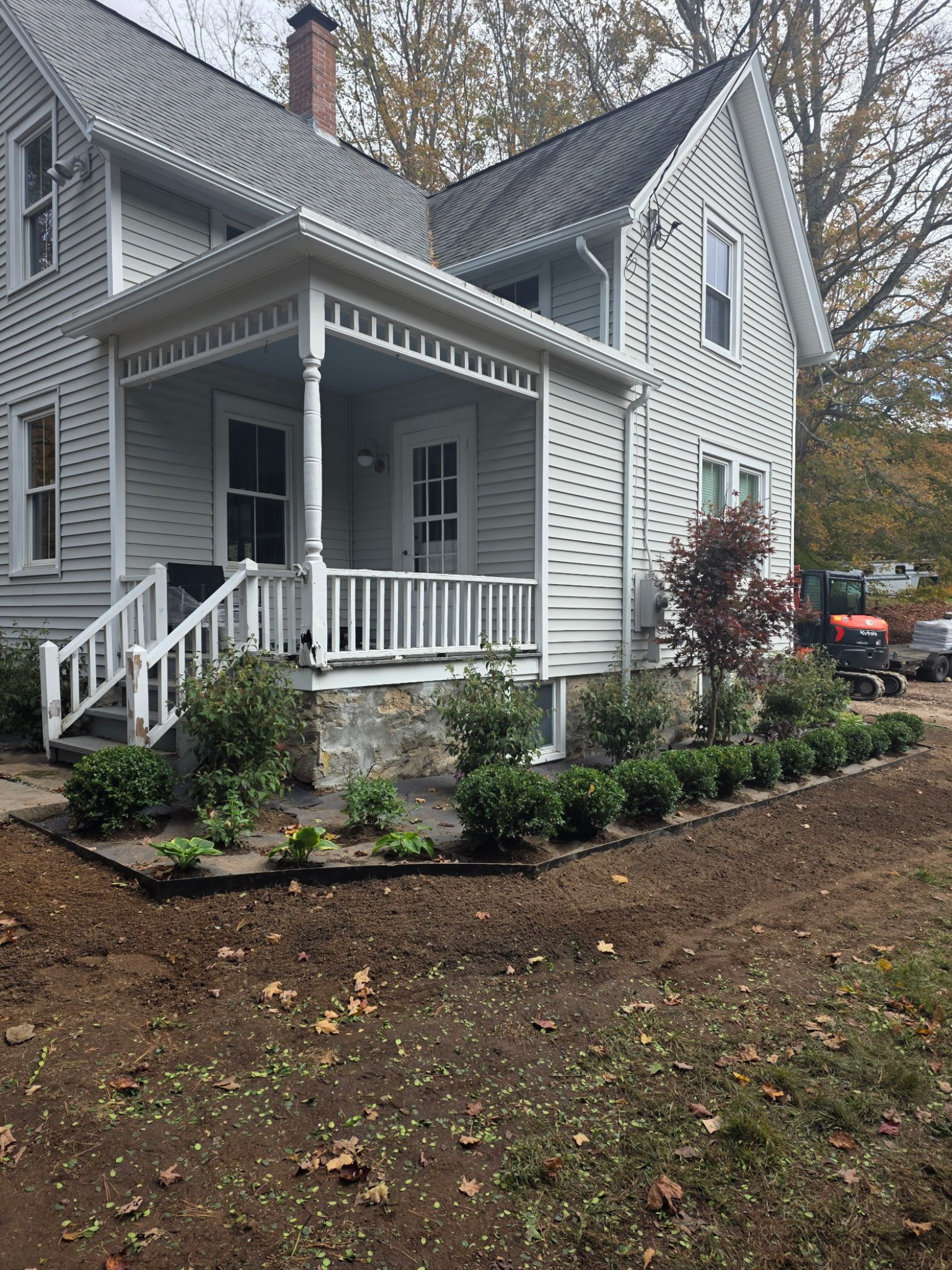 Two-story white house with a porch and new landscaping; brown dirt and green plants in the foreground.