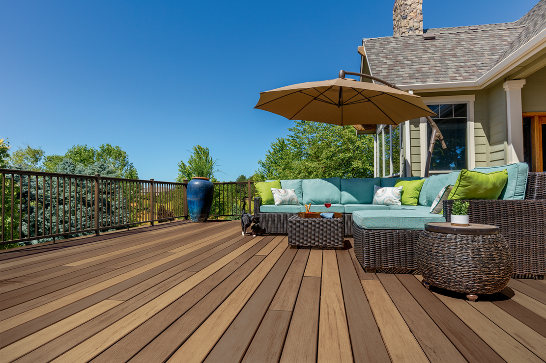 Outdoor wooden deck with wicker seating, turquoise cushions, and a tan umbrella beside a stone house.