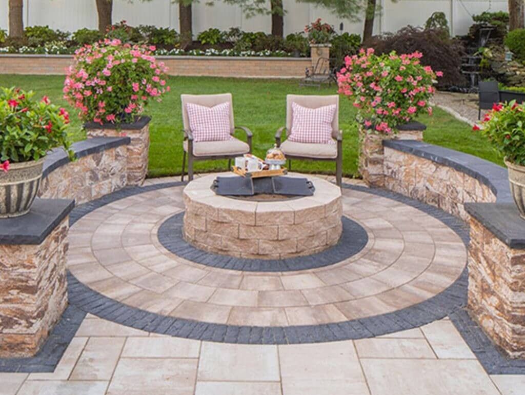 Patio seating area with two white chairs, a round stone fire pit, and pink flowering shrubs