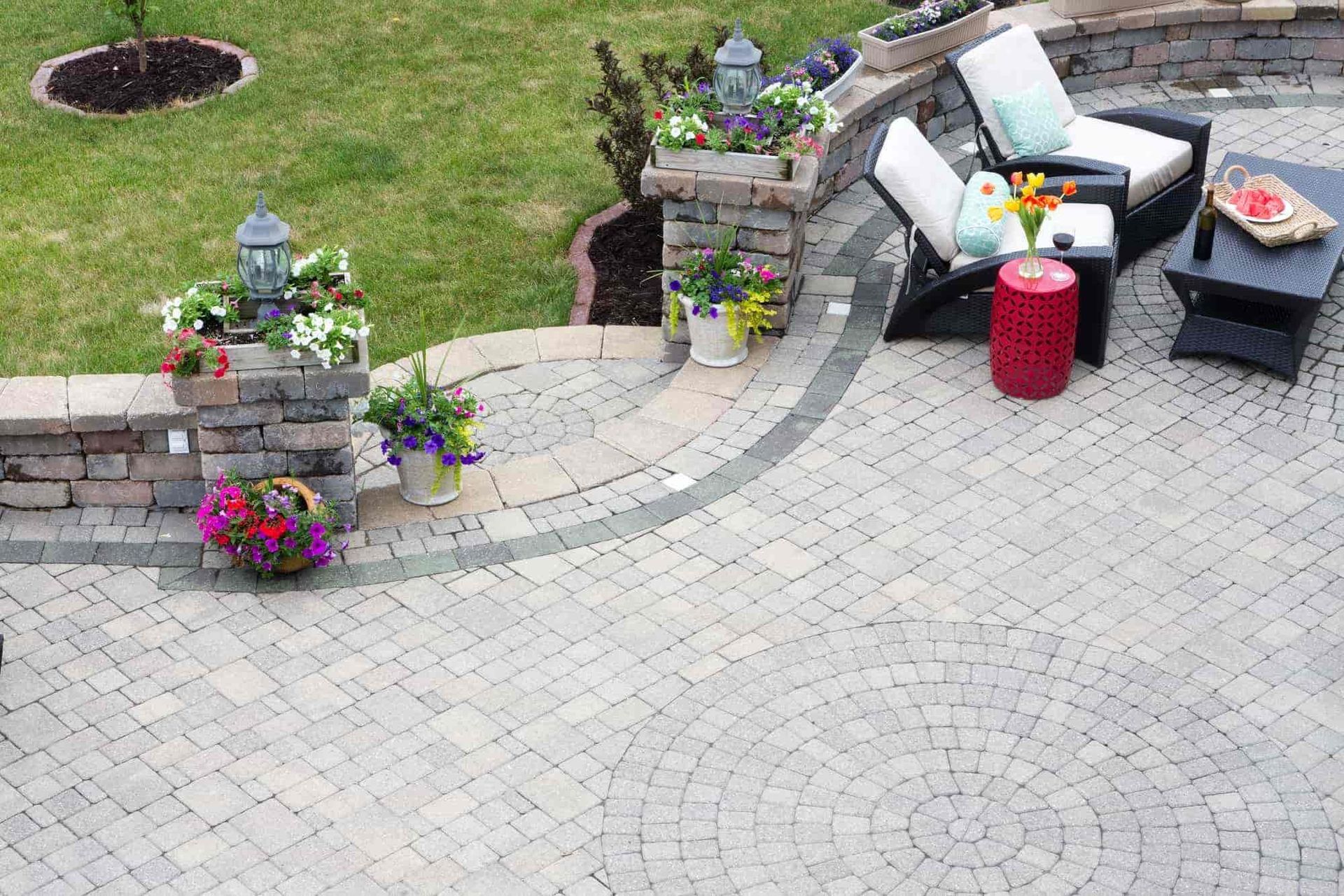 Patio with wicker chairs, red table, potted flowers, and stone planters beside a lawn