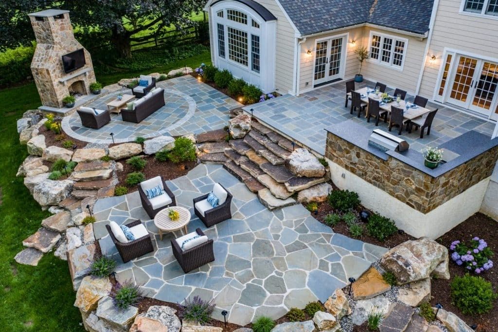 Stone patio backyard with outdoor seating, fire pit, and built-in stone fireplace beside a house
