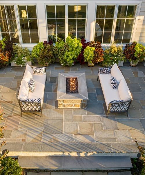 Outdoor patio with white cushioned sofas, fire pit table, and potted plants in front of windows