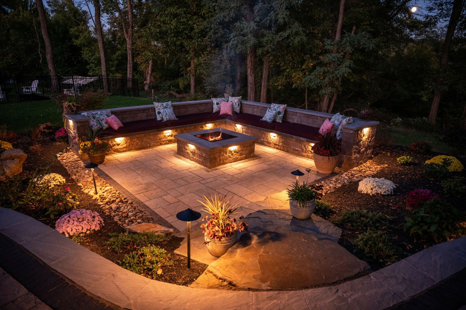 Nighttime backyard patio with a lit fire pit, curved seating, and landscaped rocks and plants