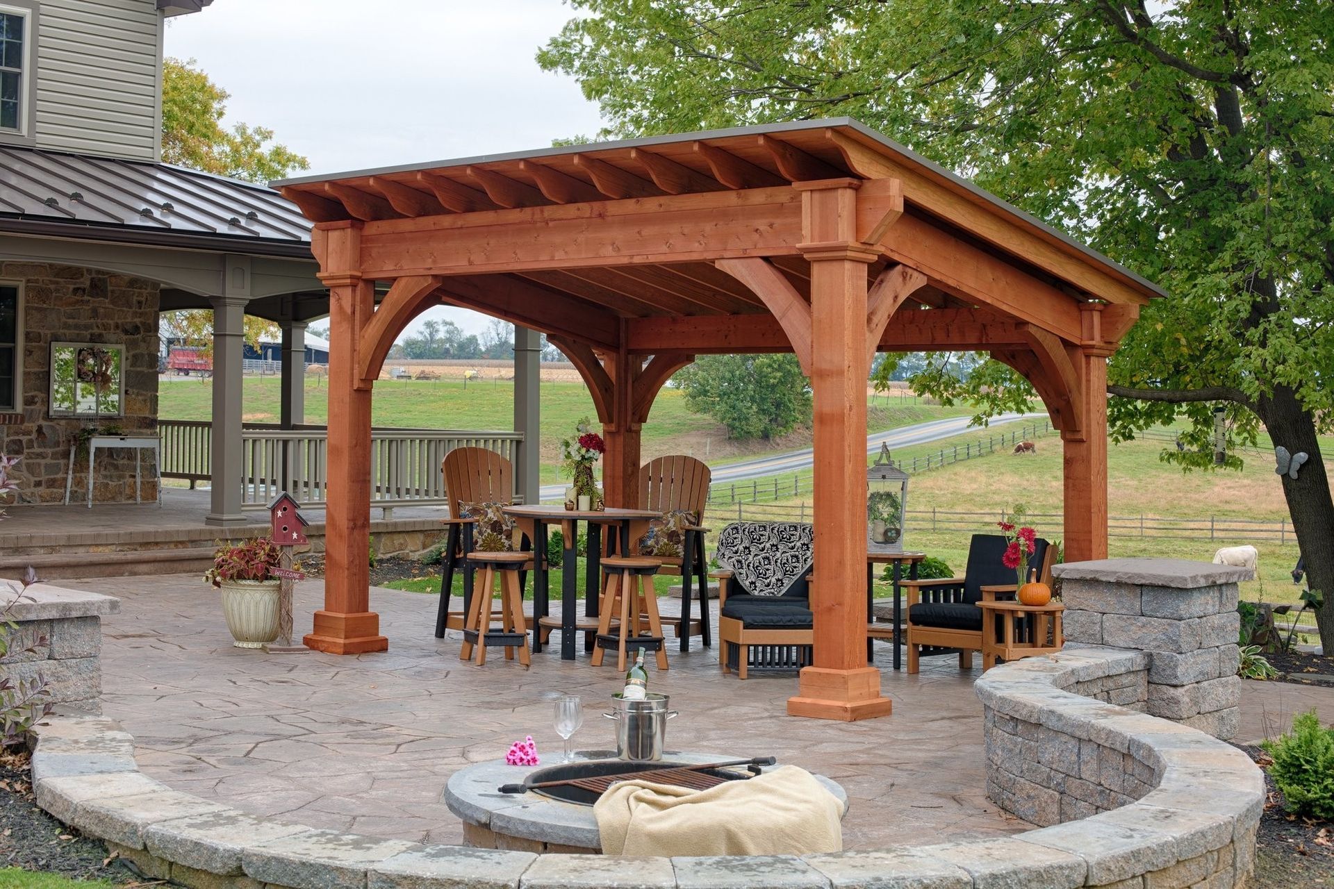Wooden pavilion with a table and chairs beside a circular stone fire pit in a backyard garden