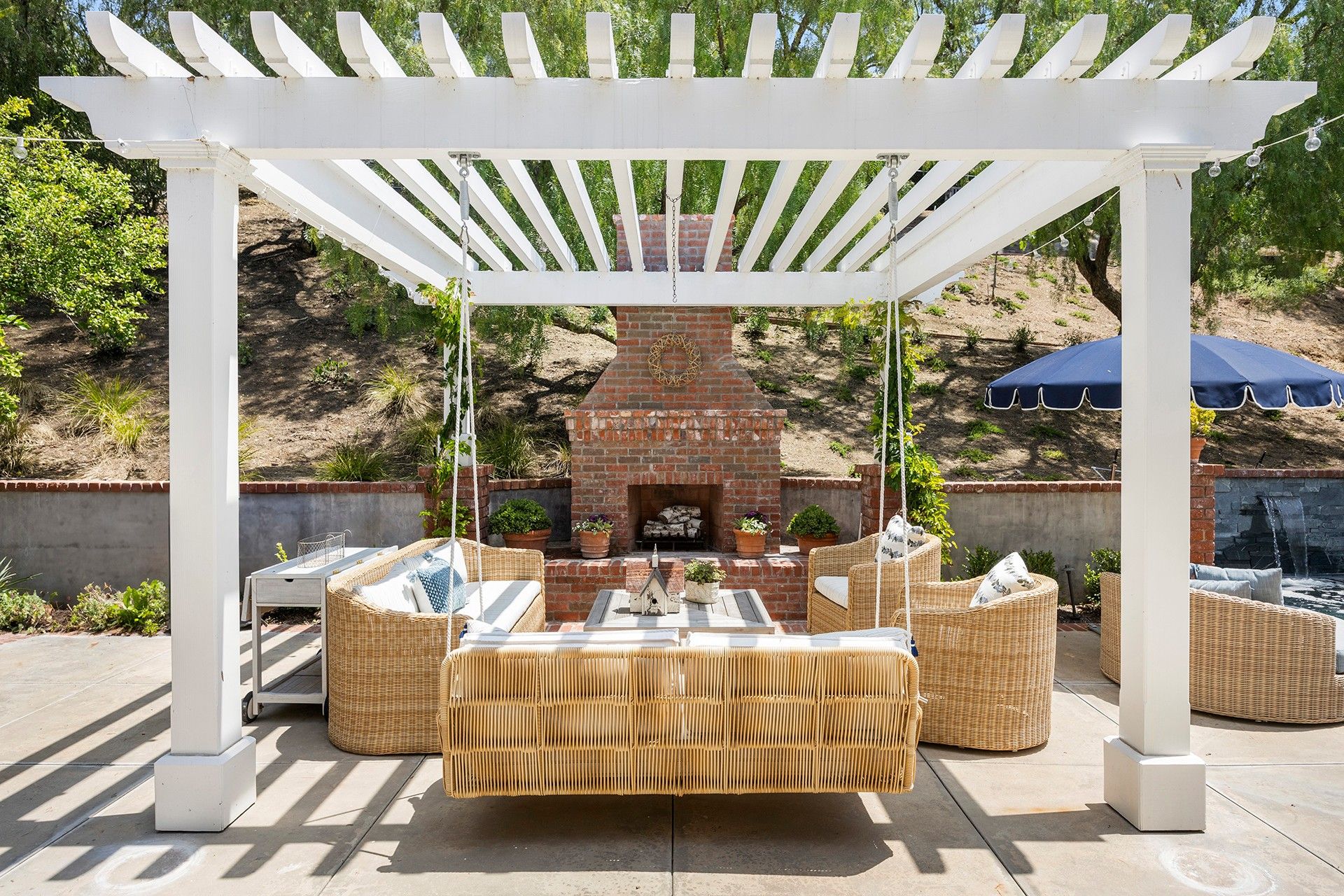 White pergola over outdoor dining area with wicker furniture and brick fireplace by a pool