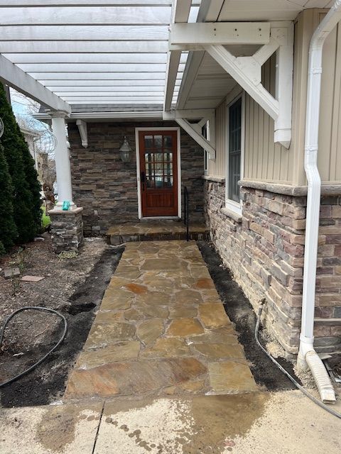 Covered stone walkway leading to a red front door beside a brick house and white awning