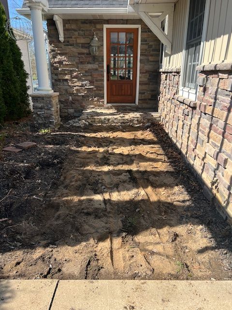 Narrow dirt walkway beside a stone-and-siding house leading to a wooden front door in sunlight