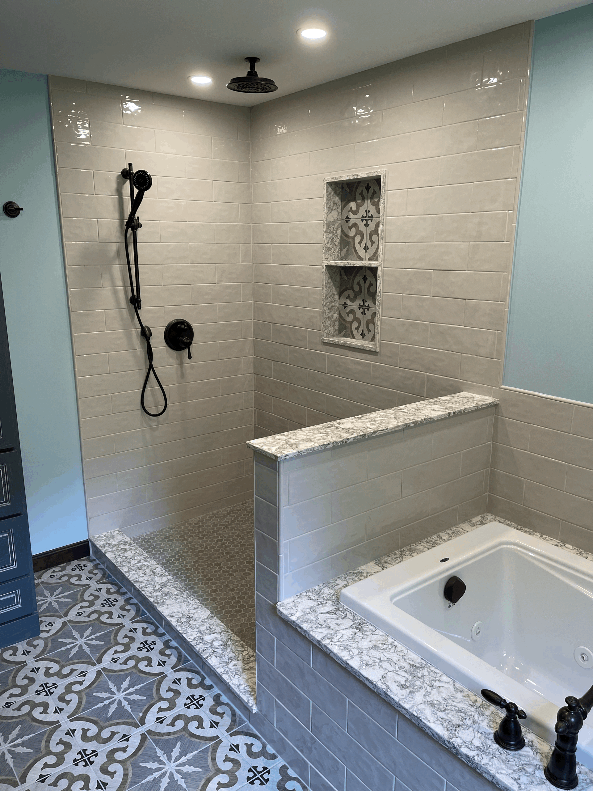 Modern bathroom with tiled shower, tub, and patterned floor. Light blue walls and black fixtures.