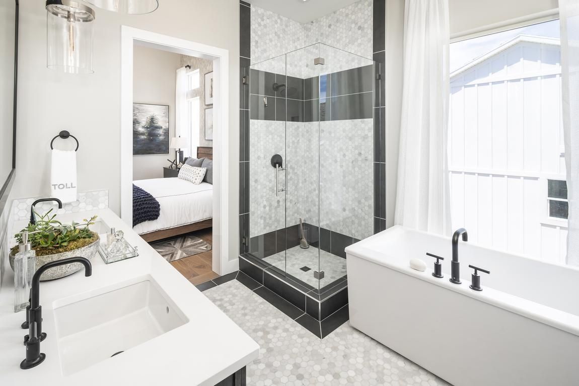 Modern bathroom with white tub, shower, vanity, and black fixtures; bedroom visible in the doorway.