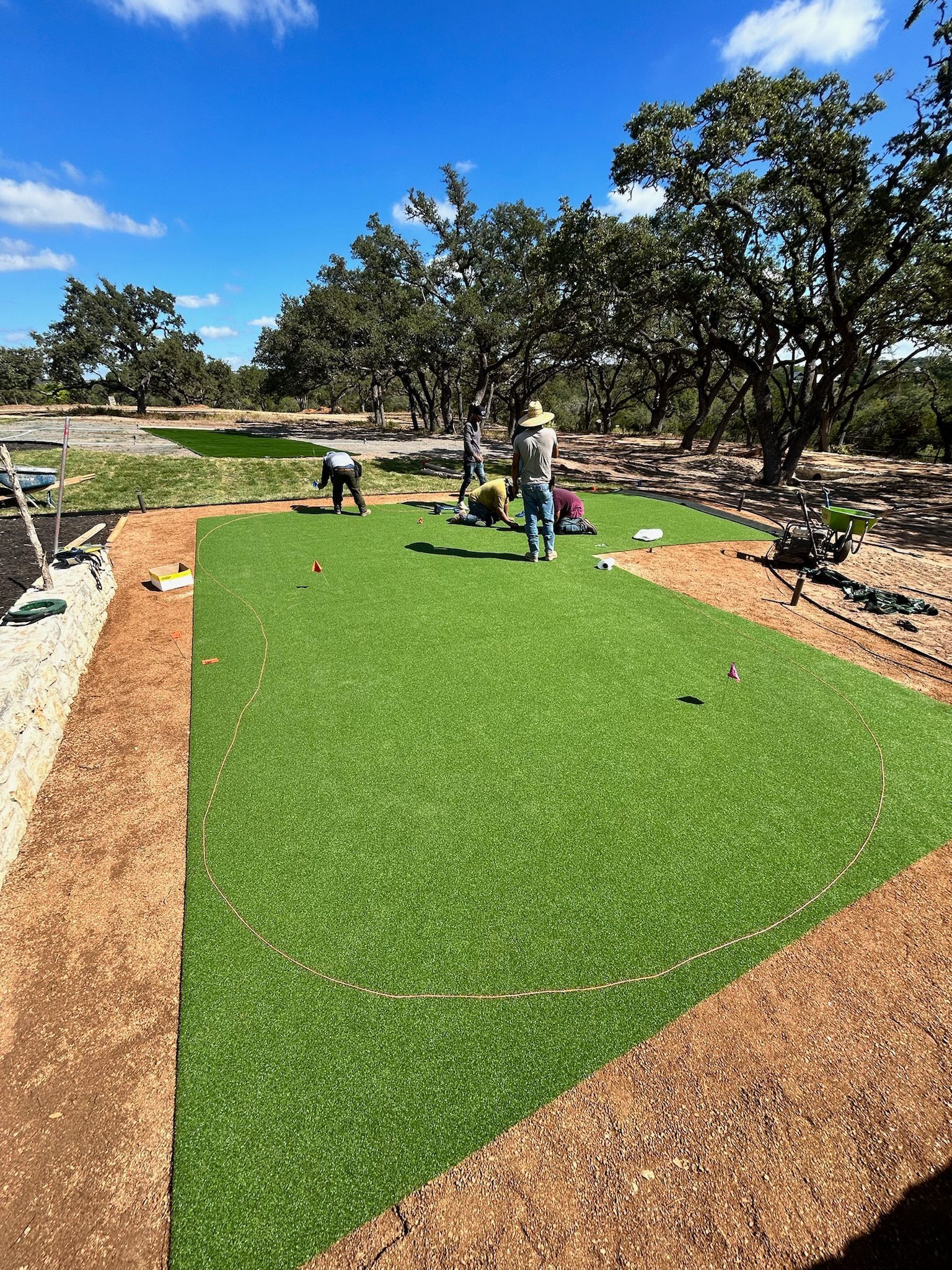People installing green artificial turf in a landscaped yard with trees and gravel paths.