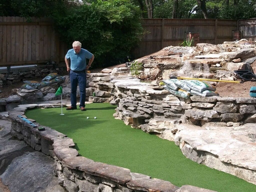A man standing on a backyard putting green beside a rocky landscape feature and a wooden fence.