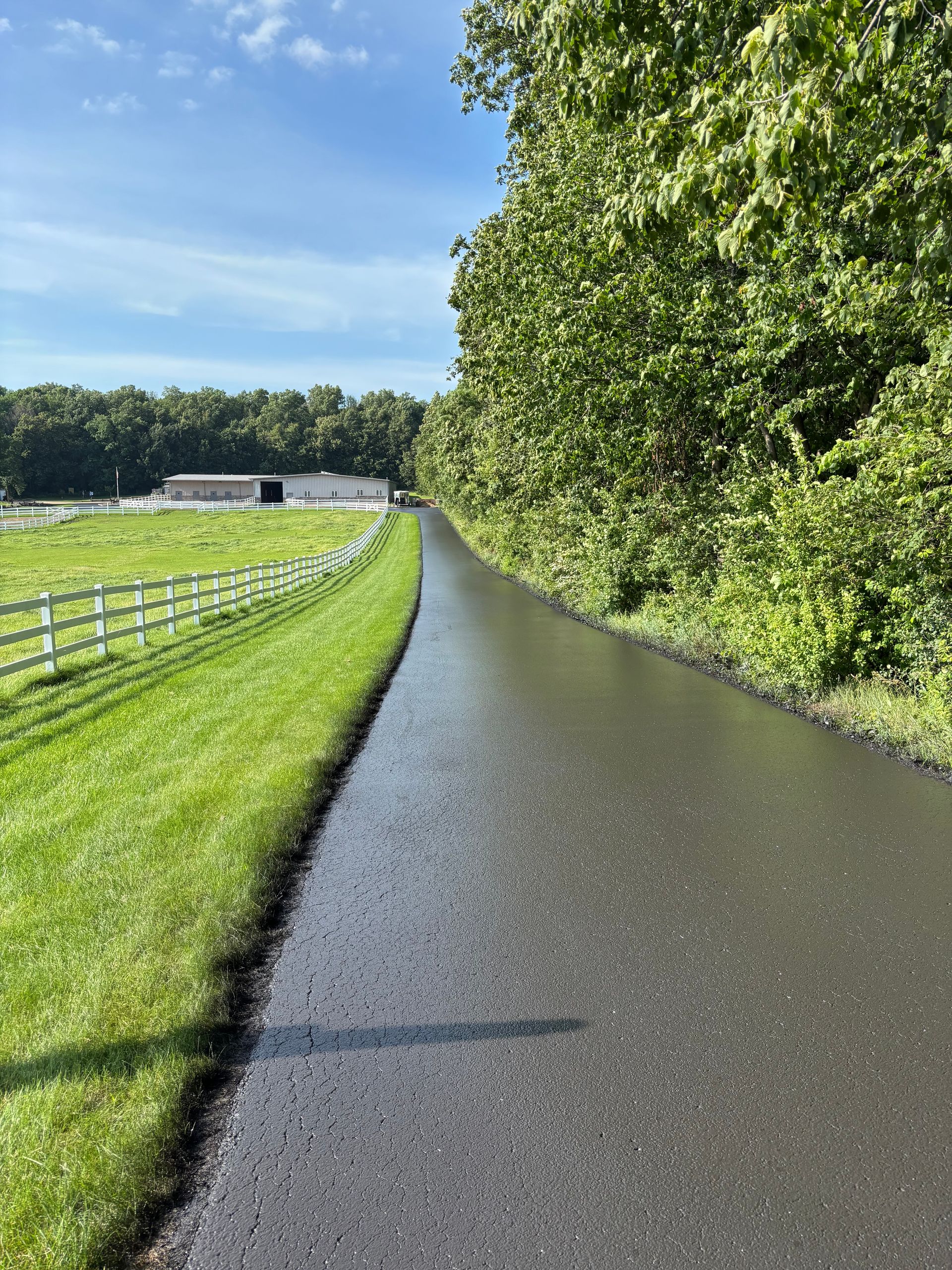 Paved road between green grass and trees, on a sunny day. White fence and barn in the distance.