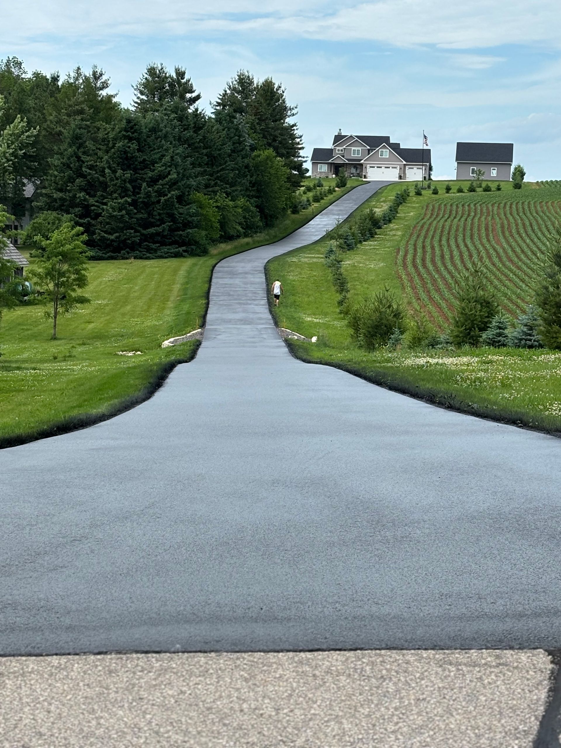 Long, freshly paved asphalt driveway leading uphill to a house and landscape with green grass and trees.