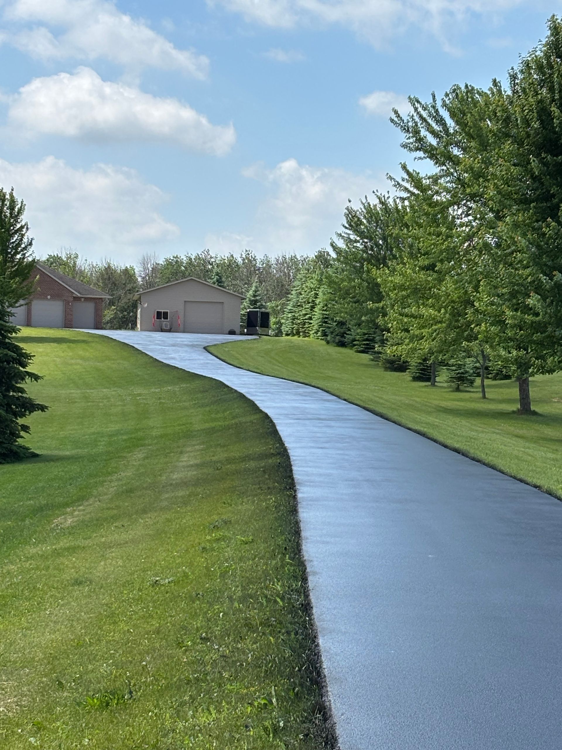 Long, black asphalt driveway curves towards two tan garages, flanked by green grass and trees under a blue sky.