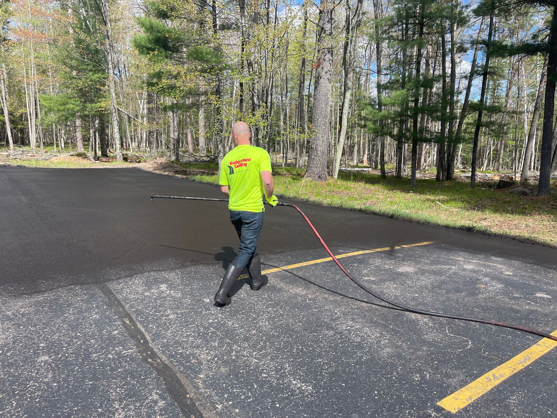 Man in neon shirt spraying asphalt pavement with a hose outdoors, trees in background.
